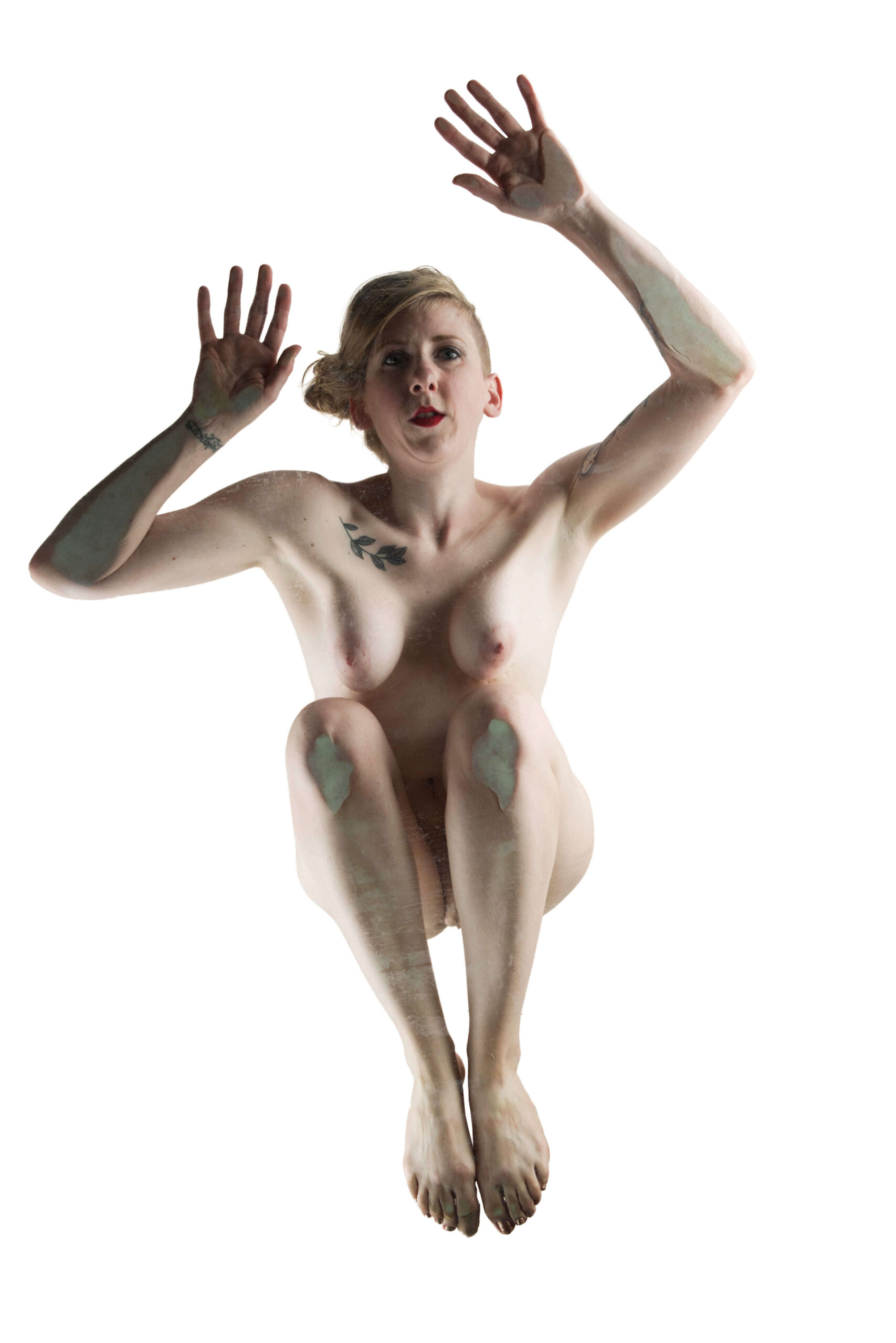 Hippy Researcher poses nude with both arms raised and knees drawn upward against a bright white background in a striking high-key fine art nude portrait.