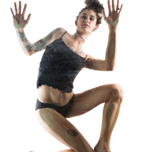 Greasyrose suspended in a dynamic airborne pose against a brilliant white background, wearing a black lace-trimmed camisole and matching underwear. Her tattooed arms extend upward with open palms, while her bent legs create a striking sense of weightless movement.
