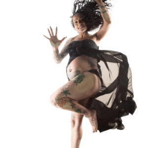 Maxi Soule poses above a glass surface with curly hair, raised hands, colorful tattoos, and flowing black fabric against a white background.