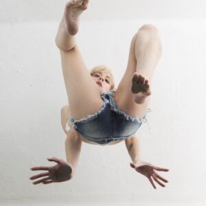 Paige Elithe Hoffman poses in frayed denim shorts with bare feet and legs raised toward the camera in a dramatic low-angle studio photograph.