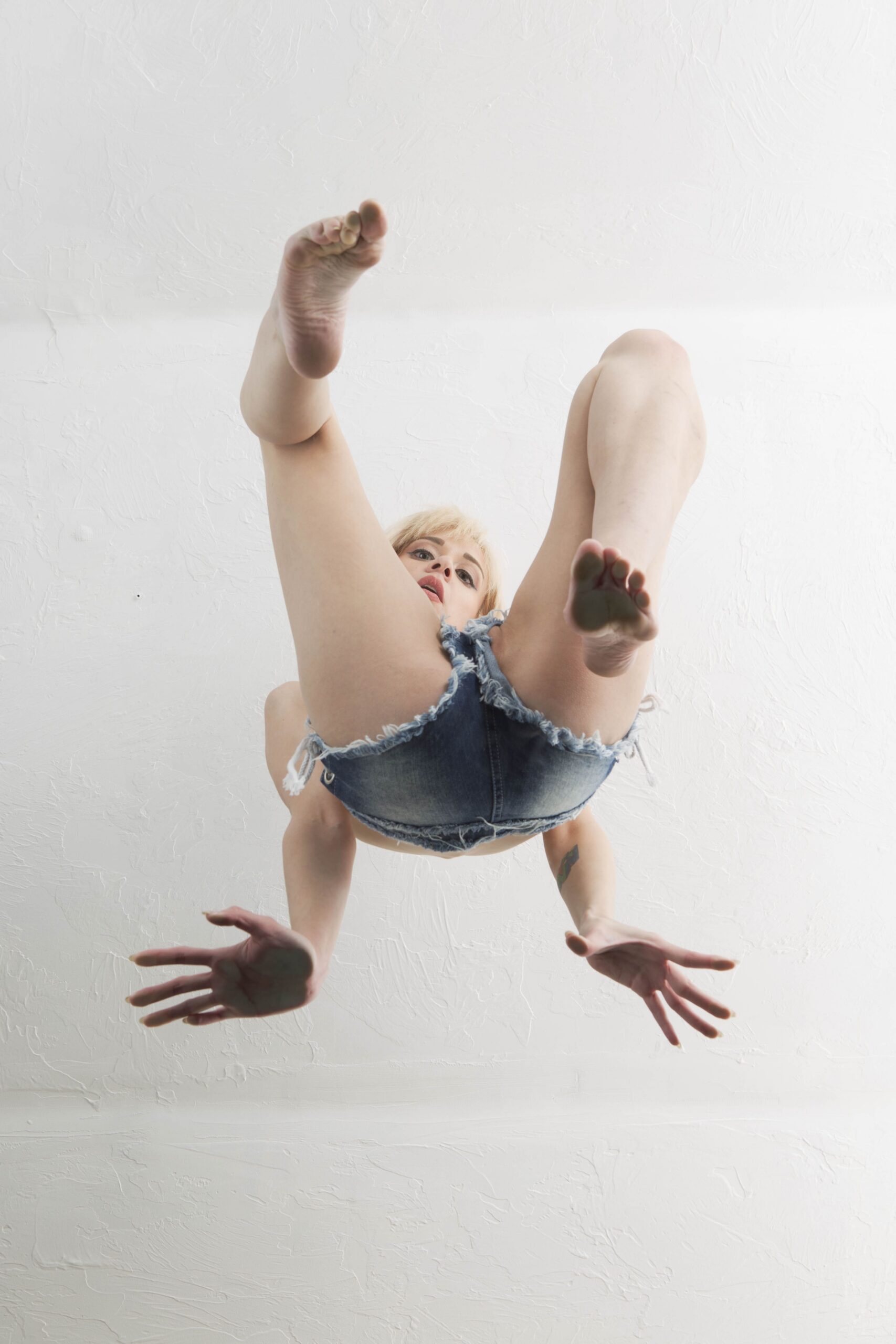 Paige Elithe Hoffman poses in frayed denim shorts with bare feet and legs raised toward the camera in a dramatic low-angle studio photograph.