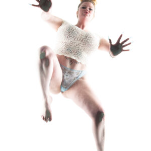 Peachesthe_real poses above a low-angle camera in a white lace crop top and embroidered lingerie, with raised hands and bent legs against a white background.