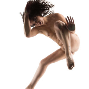Fine art nude photograph of Julia Wagner suspended in midair against a brilliant white background, with flowing curly hair and a dynamic, sculptural pose.