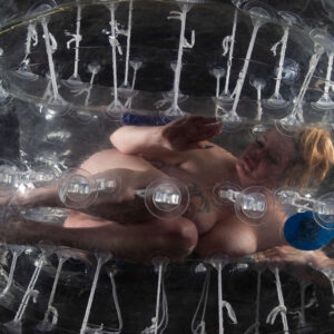 Peachesthe_real curled inside a transparent inflatable sphere surrounded by white ropes and suction cups against a black studio background.