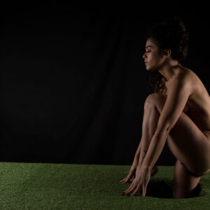 Fine art portrait of Julia Wagner crouching on a green grass-covered platform against a black studio background, with dramatic low-key lighting highlighting her figure.