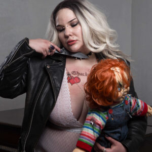 El The Belle poses in white lace lingerie and a black leather jacket, holding a red-haired Chucky doll against her body. With platinum blonde waves, striking makeup, and a knife positioned near her neck, she creates a provocative horror-inspired portrait against a muted studio background.