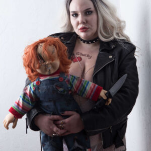 El The Belle poses in a black leather jacket, white lace lingerie, and a spiked choker while holding a sinister red-haired Chucky doll clutching a knife. Her platinum blonde waves, bold makeup, and confident expression create a striking horror-inspired portrait.