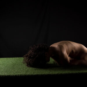 art nude photograph of Julia Wagner curled forward on a green grass-covered platform, with dramatic low-key lighting illuminating her back against a black studio background.