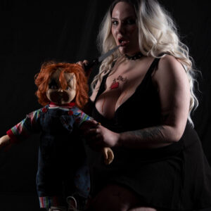 El The Belle kneels against a dark backdrop beside a sinister Chucky doll. Wearing a plunging black dress, spiked choker, and platinum blonde waves, she holds a knife near her lips, creating a provocative, horror-inspired portrait filled with danger and dark allure.