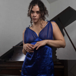Julia Wagner poses in a sapphire blue satin and sheer lace babydoll dress beside a vintage grand piano in a studio portrait.