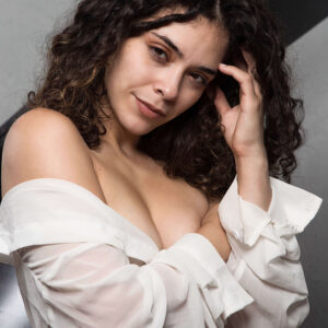 Julia Wagner poses in an off-shoulder white blouse with curly dark hair, resting her hand against her temple beside a grand piano.