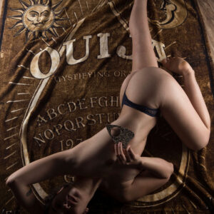 Julia Wagner reclines nude on a vintage Ouija board tapestry, holding an ornate planchette against her chest in a dramatic overhead fine art boudoir photograph.