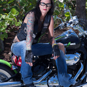 Boston sits on a black and chrome cruiser motorcycle during a Florida outdoor photography session, wearing a black leather biker vest, blue jeans, fingerless gloves, and sunglasses, showcasing her colorful tattoos.