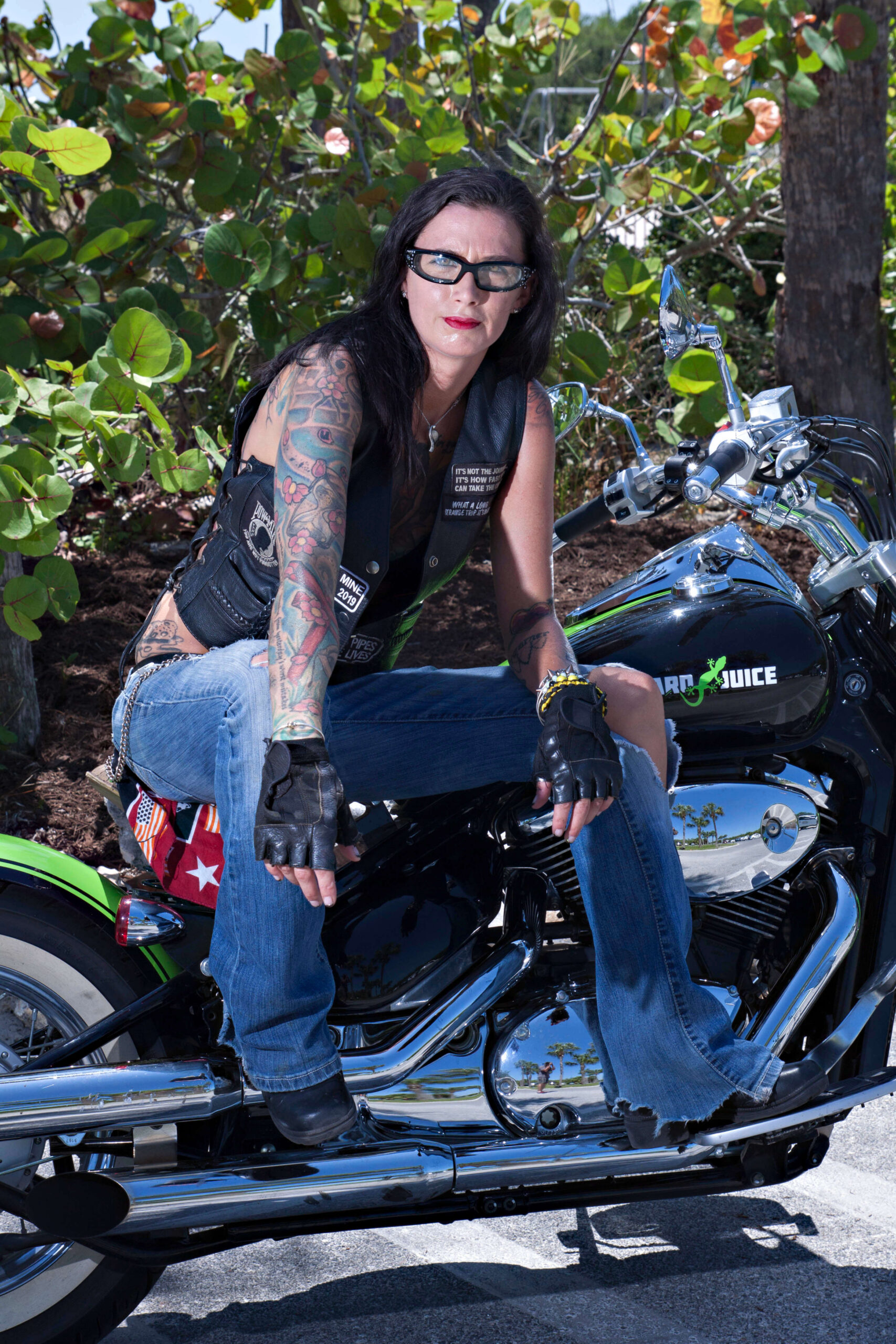 Boston sits on a black and chrome cruiser motorcycle during a Florida outdoor photography session, wearing a black leather biker vest, blue jeans, fingerless gloves, and sunglasses, showcasing her colorful tattoos.