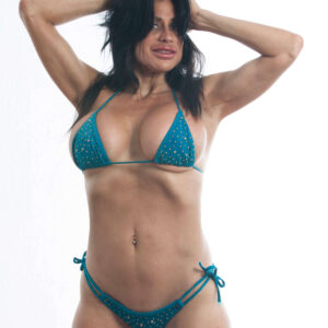 Madam Mayhem poses with both hands in her dark hair, wearing a turquoise rhinestone bikini against a bright white studio background.